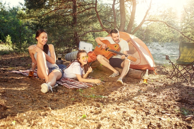 Partij, kamperen van mannen en vrouwengroep bij bos. Ze ontspanden zich en zongen een liedje tegen het groene gras. Het concept vakantie, zomer, avontuur, levensstijl, picknick