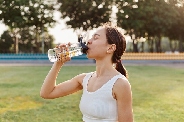 Pas donkerharig vrouwelijk drinkwater uit haar waterfles op het stadion net voordat ze begint met trainen, buiten trainen in de zomer, gezonde levensstijl.