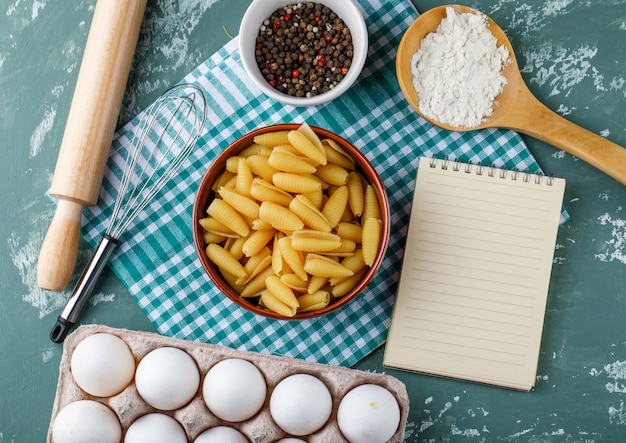 Pasta in een kom met eieren, zetmeel, peperkorrels, garde, deegroller en schrijfboek