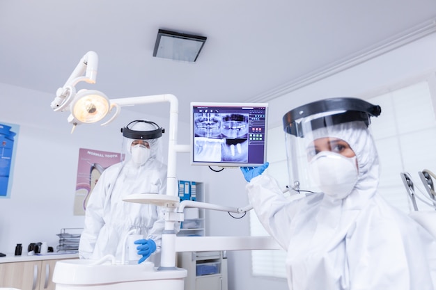 Patiënt pov van tandarts met gezichtsschild die tandheelkundige röntgenfoto's uitlegt die op de monitor wijzen. Stomatologiespecialist die een beschermend pak draagt tegen infectie met coronavirus wijzend op radiografie.