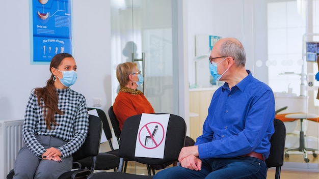 Patiënten met een beschermingsmasker praten zittend op stoelen en houden sociale afstand in de stomatologische kliniek, wachtend op arts tijdens coronavirus. Concept van nieuw normaal tandartsbezoek bij covid19-uitbraak