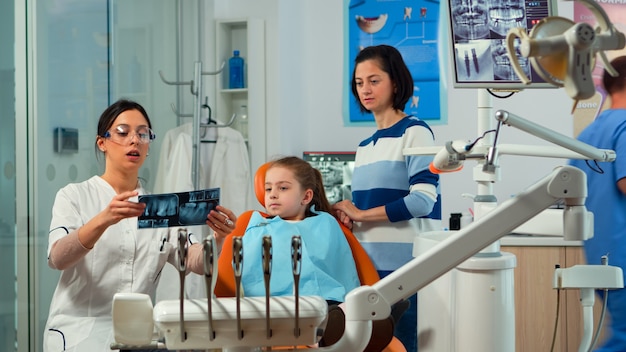 Pediatrische vrouw tandarts die kleine meisjespatiënt behandelt in een modern stomatologisch tandheelkundig kantoor met tandenröntgenfoto's die uitleggen aan de tandheelkundige interventie van de moeder. tandarts bezoeken met kinderen.