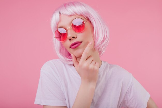 Peinzend charmant meisje met roze haar opzoeken terwijl ze poseren op pastel achtergrond Zorgeloze Europese vrouw in zonnebril en periwig chillen tijdens studio fotoshoot