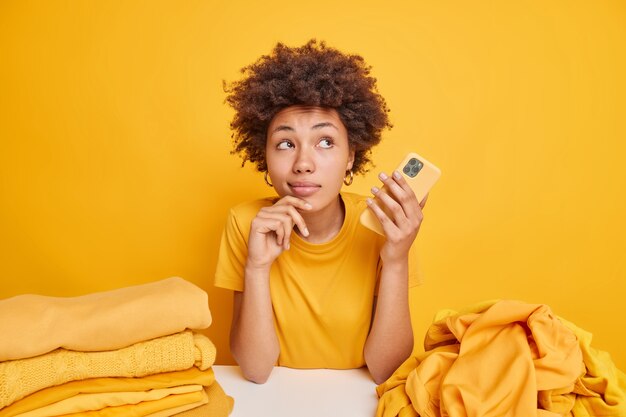 Peinzende Afro-Amerikaanse vrouw heeft een dromerige uitdrukking met een moderne mobiele telefoon aan tafel met stapels kleren geïsoleerd over een gele muur, bezig met het vouwen van de was. Kleding en huishouden