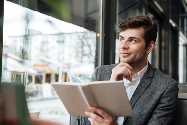 Peinzende het glimlachen zakenmanzitting door de lijst in koffie met laptop computer terwijl het houden van boek en weg het kijken