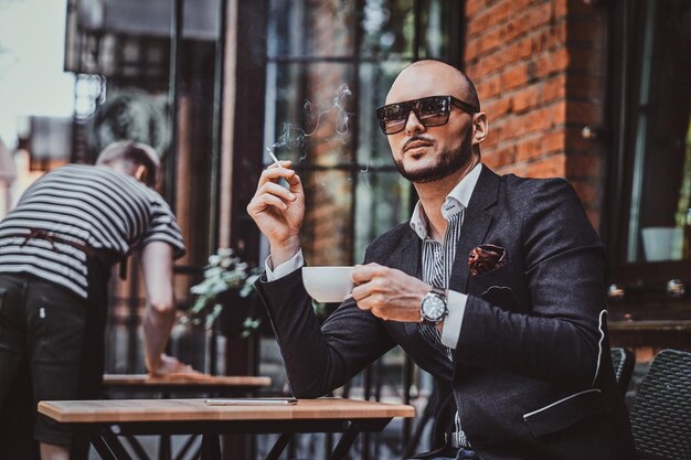 Peinzende moderne man rookt een sigaret buiten de coffeeshop terwijl hij zijn kopje koffie drinkt.