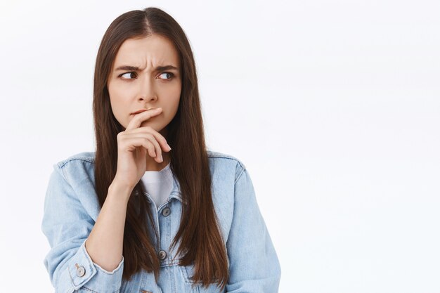 Peinzende, serieus ogende gefocuste blanke vrouw in spijkerjasje, lip attent aanraken, intens fronsen en zijwaarts kijken als denken, lastige gedachten hebben, witte achtergrond