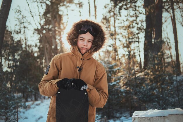 Peinzende tiener in kap poseert voor fotograaf met zijn snowboard in het winterbos.