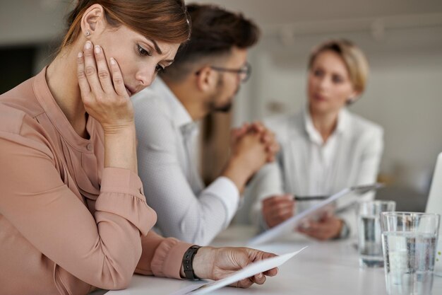 Peinzende vrouw die spuitplannen onderzoekt terwijl haar man met een makelaar op kantoor praat