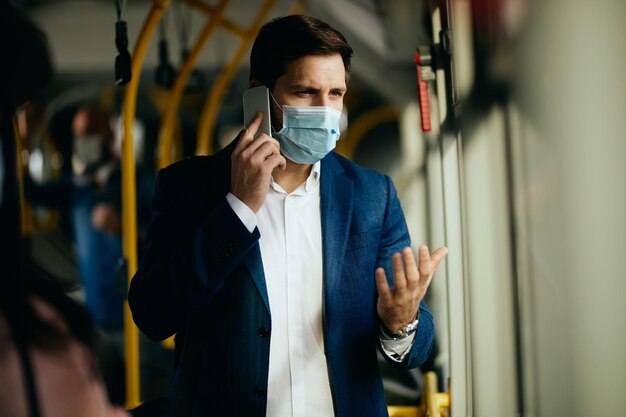 Peinzende zakenman met gezichtsmasker die aan de telefoon praat tijdens het woon-werkverkeer met de bus