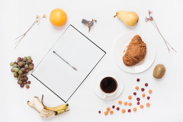 Pen op een open notitieboekje met koffiekop; croissant en veel fruit op witte achtergrond