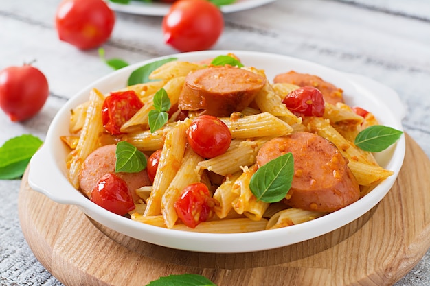 Penne pasta met tomatensaus met worst, tomaten, groene basilicum ingericht in een koekenpan op een houten tafel