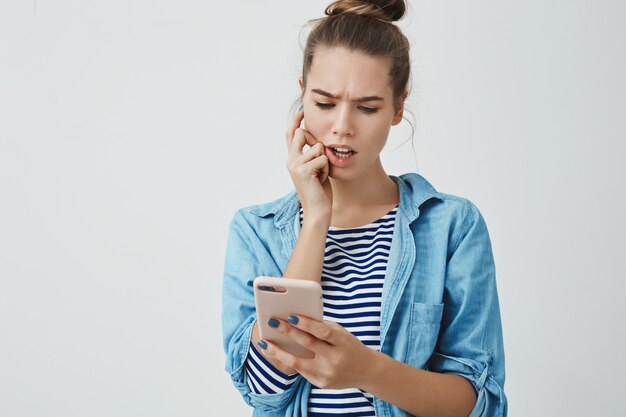 perplex verontruste schattige jonge 25s-vrouw die rekeningenlijst online controleert, fronsen verontruste bijtende vinger op zoek naar smartphone bezorgde nadenkende, berekenende geest