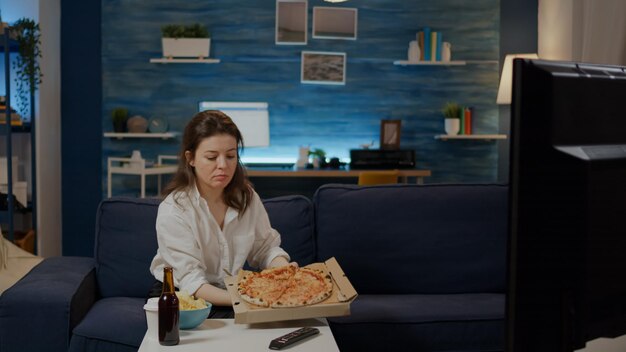 Persoon die een doos pizza van de bezorger op tafel in de woonkamer brengt. Jonge vrouw met fastfood en alcoholische drank zittend op de bank televisie kijken. Volwassene bestellen afhaalmaaltijd