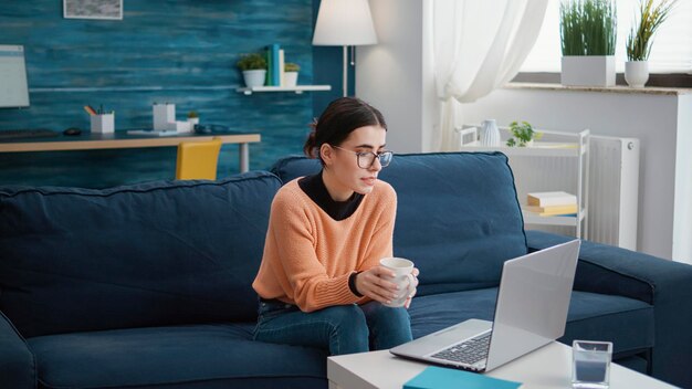 Persoon die een gesprek op afstand bijwoont tijdens een videogesprek, praat met de leraar op een teleconferentie voor een online klasles. Vrouw zittend op de bank en met behulp van laptop met videoconferentie.