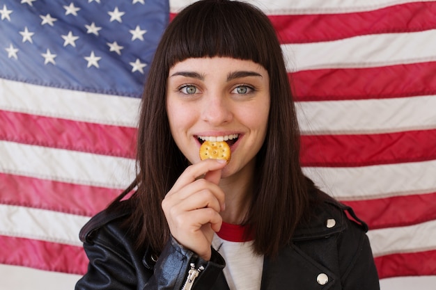 Petite vrouwelijke vrouw of tiener in trendy hipster outfit eet poses met knapperige pindakaas koekjescracker, typische traditionele Amerikaanse snack met usa vlag