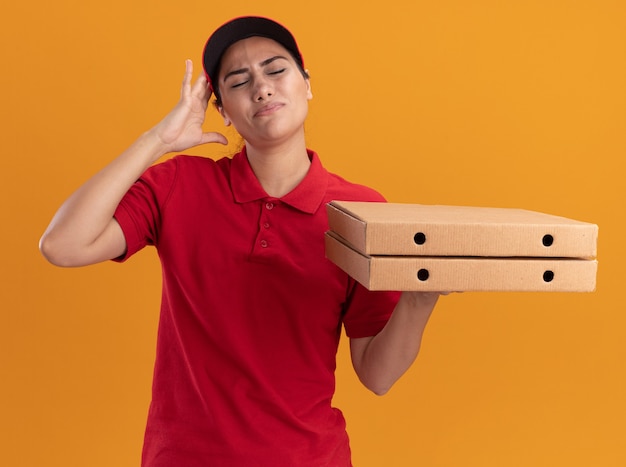 Pijn met gesloten ogen jong levering meisje dragen uniform en pet houden pizzadozen hand op het hoofd zetten geïsoleerd op oranje muur