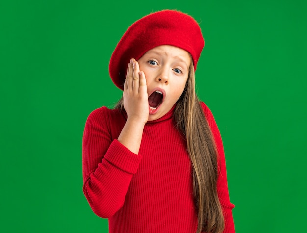 Pijnlijk klein blond meisje met een rode baret die naar de voorkant kijkt en de hand op de kin houdt met open mond geïsoleerd op een groene muur met kopieerruimte