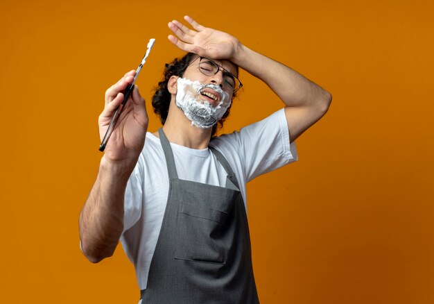 Pijnlijke jonge blanke mannelijke kapper met bril en golvende haarband in uniform met scheermes hand op het hoofd zetten met scheerschuim op zijn gezicht met gesloten ogen