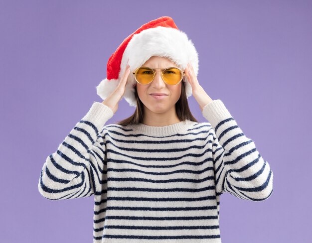 pijnlijke jonge blanke meisje in zonnebril met kerstmuts legt handen op het hoofd geïsoleerd op paarse achtergrond met kopie ruimte