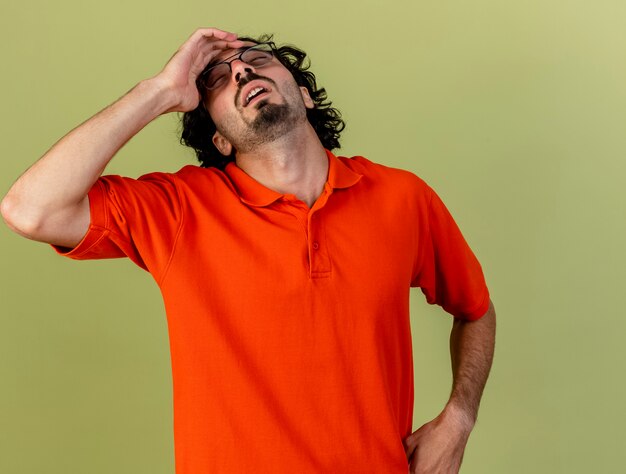 Pijnlijke jonge blanke zieke man met bril hoofd aan te raken met gesloten ogen geïsoleerd op olijfgroene achtergrond met kopie ruimte