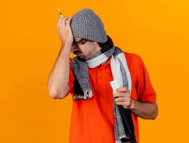 Pijnlijke jonge blanke zieke man met bril, muts en sjaal met spuit en pakje medische tabletten hand op het hoofd houden met gesloten ogen geïsoleerd op oranje muur met kopie ruimte
