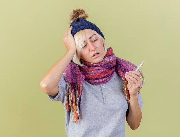 Pijnlijke jonge blonde zieke Slavische vrouw met winter muts en sjaal legt hand op het hoofd en houdt thermometer geïsoleerd op olijfgroene muur met kopie ruimte