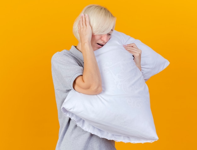 Gratis foto pijnlijke jonge blonde zieke vrouw legt hand op het hoofd en knuffels kussen geïsoleerd op oranje muur