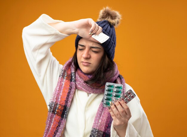 Pijnlijke jonge Kaukasische ziek meisje dragen gewaad winter muts en sjaal met verpakkingen van capsules en tabletten hand op het hoofd houden met gesloten ogen geïsoleerd op oranje muur