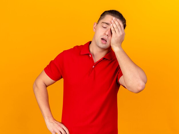 Pijnlijke jonge knappe blonde zieke man aanraken hoofd houden hand op taille met hoofdpijn met gesloten ogen geïsoleerd op oranje muur