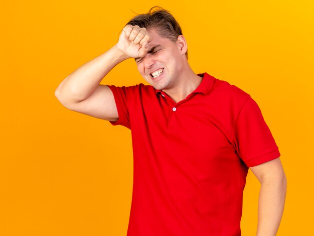 Pijnlijke jonge knappe blonde zieke man die hand op voorhoofd houdt die aan hoofdpijn lijden met gesloten ogen die op oranje muur wordt geïsoleerd