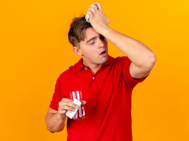 Pijnlijke jonge knappe blonde zieke man met pakje medische tabletten glas water en servet hoofd aanraken met hoofdpijn met gesloten ogen geïsoleerd op oranje muur