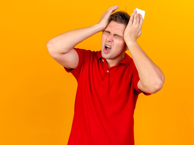 Pijnlijke jonge knappe blonde zieke man met servet handen op het hoofd houden die lijden aan hoofdpijn met gesloten ogen geïsoleerd op oranje muur met kopie ruimte