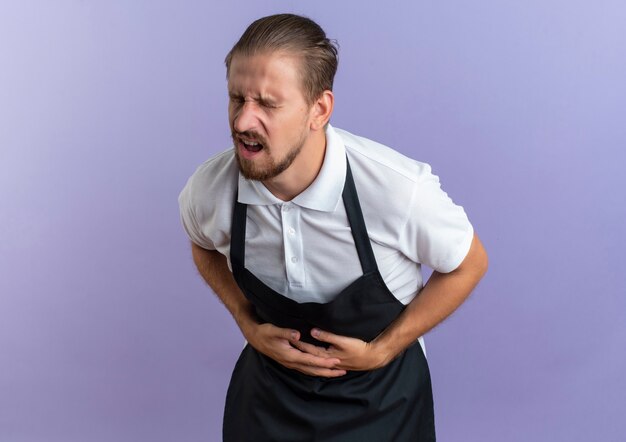 Pijnlijke jonge knappe kapper die uniform draagt ?? die zijn buik houdt die aan pijn lijden met gesloten ogen die op paars met exemplaarruimte wordt geïsoleerd