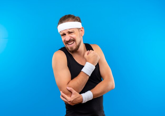 Pijnlijke jonge knappe sportieve man met hoofdband en polsbandjes die de vuist balt en zijn elleboog geïsoleerd op blauwe muur met kopieerruimte houdt