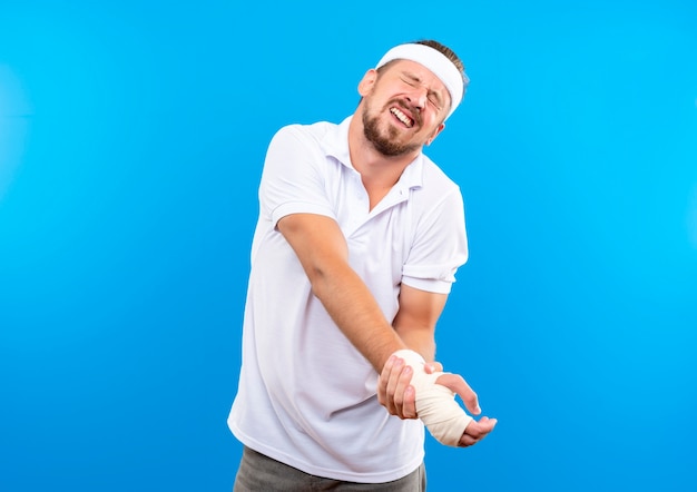 Pijnlijke jonge knappe sportieve man met hoofdband en polsbandjes met zijn gewonde pols omwikkeld met verband met gesloten ogen geïsoleerd op blauwe muur met kopieerruimte