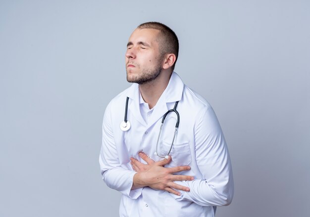 Pijnlijke jonge mannelijke arts die medische mantel en stethoscoop om zijn nek draagt, die zijn buik met gesloten ogen houdt die op wit met exemplaarruimte worden geïsoleerd