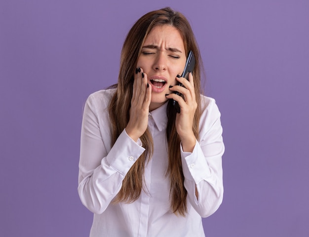 Pijnlijke jonge, mooie blanke meid legt hand op gezicht en praat over telefoon geïsoleerd op paarse muur met kopieerruimte