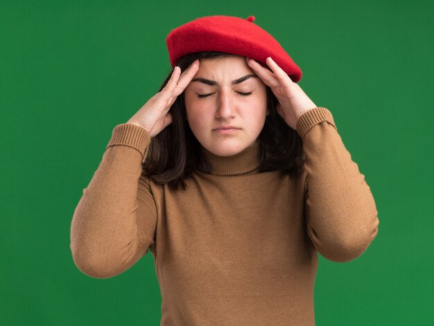 Gratis foto pijnlijke jonge mooie blanke meisje met baret hoed legt handen op voorhoofd op groen