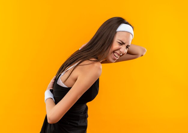 Pijnlijke jonge mooie sportieve meid met hoofdband en polsband die handen op haar rug legt in profielweergave geïsoleerd op oranje muur