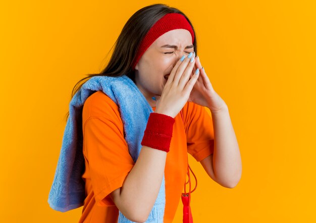 Pijnlijke jonge sportieve vrouw die hoofdband en polsbandjes met handdoek en springtouw op schouders draagt die neus met gesloten ogen houdt