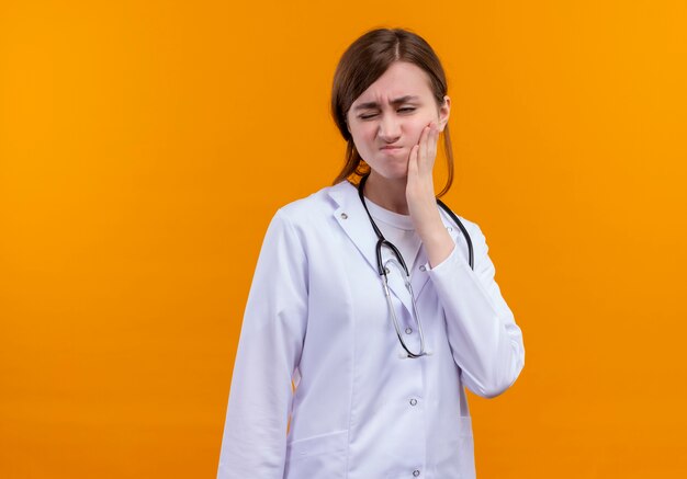 Pijnlijke jonge vrouwelijke arts die medische gewaad en stethoscoop draagt die hand op Wang zetten die aan kiespijn lijden op geïsoleerde oranje ruimte met exemplaarruimte