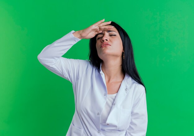 Pijnlijke jonge vrouwelijke arts dragen medische gewaad hand zetten voorhoofd lijden aan hoofdpijn met gesloten ogen geïsoleerd op groene muur met kopie ruimte