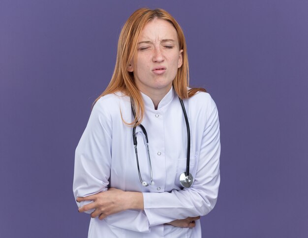 Pijnlijke jonge vrouwelijke gemberdokter die medische mantel en stethoscoop draagt en hand op de buik houdt met gesloten ogen geïsoleerd op paarse muur met kopieerruimte
