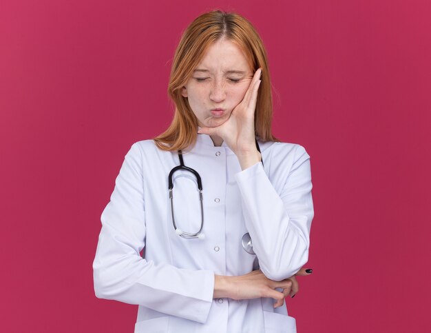 Pijnlijke jonge vrouwelijke gemberdokter die medische mantel en stethoscoop draagt en hand op het gezicht houdt, lippen tuitend met gesloten ogen met kiespijn