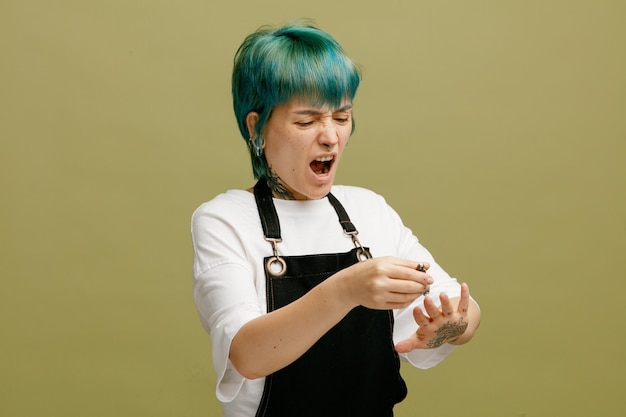 Pijnlijke jonge vrouwelijke kapper in uniform die haar nagels knipt met nagelknipper geïsoleerd op olijfgroene achtergrond