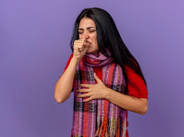 Pijnlijke jonge zieke vrouw die sjaal draagt die hand op borst met gesloten ogen zet, hoestend die vuist dichtbij mond houdt die op purpere muur wordt geïsoleerd