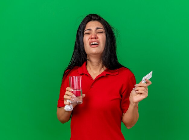 Pijnlijke jonge zieke vrouw met pak tabletten glas water en servet met gesloten ogen geïsoleerd op groene muur