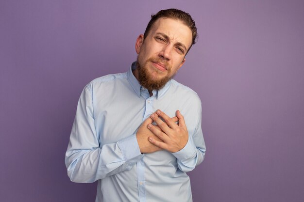 Pijnlijke knappe blonde man legt handen op hart kijken naar voorzijde geïsoleerd op paarse muur