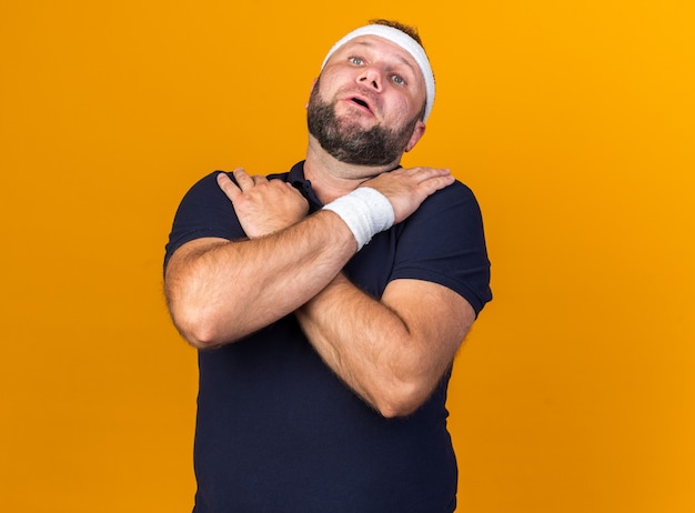 pijnlijke volwassen Slavische sportieve man met hoofdband en polsbandjes legt handen op zijn schouders geïsoleerd op oranje muur met kopieerruimte
